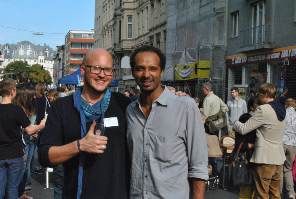 Zwei lachende Männer auf der BLOCK PARTY in der Lindenstraße, einer hebt den Daumen; im Hintergrund feiern Nachbar*innen auf der Straße.
