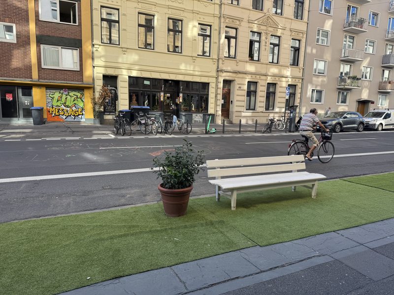 Eine weiße Sitzbank und ein Pflanzkübel mit einem grünen Strauch stehen auf einem Streifen Kunstrasen am Rand des Gehwegs. Dahinter verläuft der Radfahrstreifen, auf dem ein Radfahrer fährt.